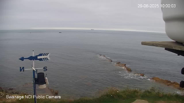 A view of the sea with a rocky breakwater extending into the water. The sky is overcast, and there are a few small boats visible in the distance. A weather vane with an arrow and directional markers (N, S) is in the foreground, along with part of a white container or structure on the right. The timestamp shows the date and time as June 29, 2025, at 10:00:03.