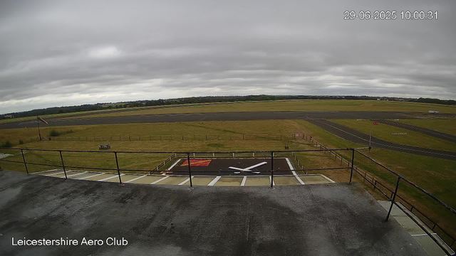 An overcast view of an airfield runway from a webcam positioned high above, showing a small helipad marked with a white "X" in the foreground, surrounded by grass and safety barriers. The runway extends into the distance with some taxiways and movement areas visible, under a cloudy sky. The location is identified as Leicestershire Aero Club.
