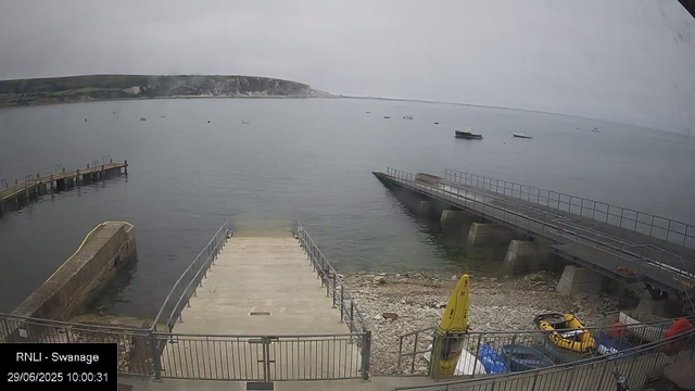 A view of a small harbor on a cloudy day. There are two piers extending into calm water, one on the left and one on the right. Several small boats are scattered across the water in the distance. In the foreground, there is a concrete dock with metal railings, and a yellow caution sign is placed near the edge. At the bottom right, there's roughly parked equipment, including a yellow inflatable boat, a blue bottle, and other miscellaneous items. The background features a sloped shoreline with green patches and white chalk cliffs under a gray, overcast sky.