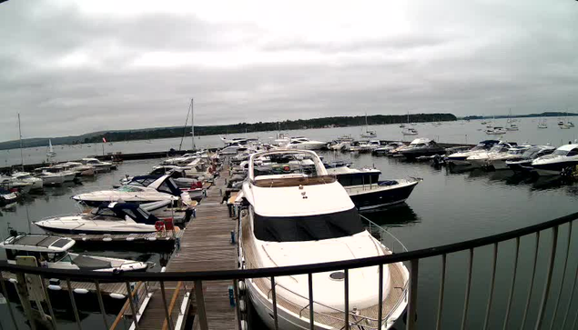 A marina filled with various boats and yachts docked along wooden piers, with calm water and an overcast sky in the background.