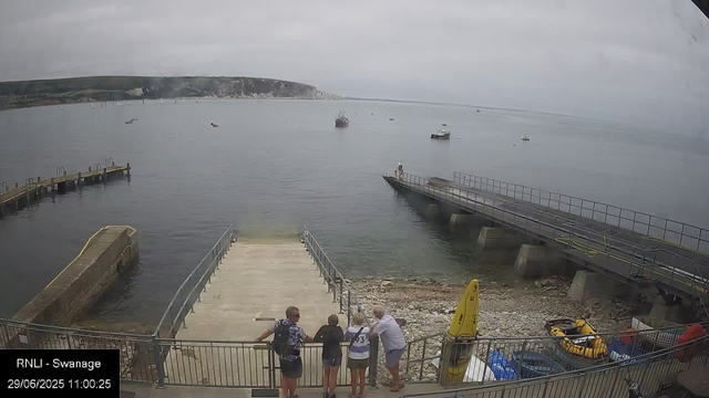 A group of five people is standing on a concrete and rocky dock by the water, engaging in conversation. The dock extends into a calm harbor with several boats anchored in the background. The scene is overcast with gray skies, and there is a hilly coastline visible in the distance. A yellow inflatable boat and some equipment are stored nearby. The timestamp indicates it is 11:00 AM on June 29, 2023.