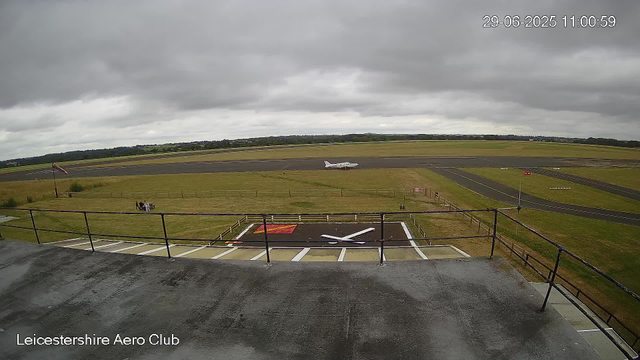 A view from a webcam at Leicestershire Aero Club shows an overcast sky with dark clouds. In the foreground, there is a helicopter landing pad with a white helicopter parked on the runway behind a black metal railing. To the left, a windsock indicates wind direction. A small bench with two people sitting on it is visible on the grassy area near the pad. Beyond the grassy area, there are taxiways and runways with a small white airplane on one of them. The horizon features a flat landscape with fields and patches of trees. The timestamp in the top right corner reads "29-06-2025 11:00:59".