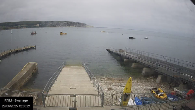 A view of a harbor with a concrete pier extending into calm water, surrounded by boats and sailboats in the distance. There is a grassy cliffside in the background under a cloudy sky. In the foreground, there are yellow marine safety equipment and a small boat, with a metal fence enclosing the area. The timestamp reads "29/06/2025 12:00:21."