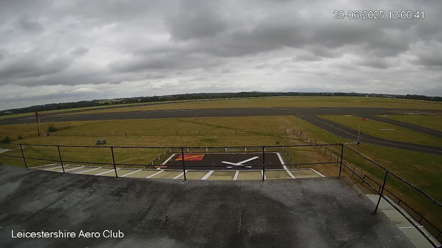 A view from a webcam showing an aircraft parking and runway area at Leicestershire Aero Club. The image features a cloudy sky, an open grassy field, and distant runways. In the foreground, there is a helicopter landing pad with a white helicopter silhouette and a red marking, surrounded by a black fence and pavement area. A small sign with the number 5 is visible near the grassy area. The timestamp shows the date and time as June 29, 2025, at 12:00:41.