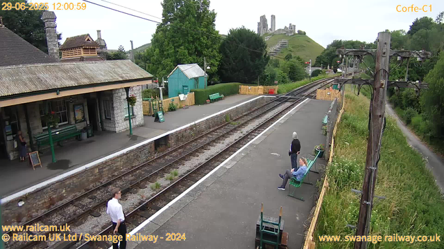 A small railway station platform with three people sitting on green benches and one person standing. The station building, made of stone with a tiled roof, is on the left side. There are flower baskets hanging outside the building. Nearby, a small teal-colored shed and a yellow fence are visible. In the background, a hill with a castle ruin on top can be seen. There are tall trees and greenery surrounding the area. An overhead power line and a large wooden pole are also present along the platform. The scene appears to be during daytime with overcast weather.