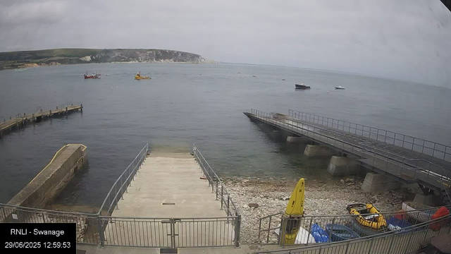 A view of a harbor with a concrete dock extending into the water, surrounded by metal railings. Several small boats are anchored in the water, and foggy cliffs can be seen in the distance. The sky is overcast with gray clouds. In the foreground, there is a yellow safety sign, and some equipment, including a yellow inflatable boat, is visible near the bottom right corner. The image has a timestamp of "29/06/2025 12:59:53" and is labeled "RNLI - Swanage."