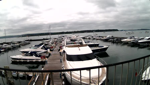 A marina filled with numerous docked boats and yachts, with a wooden pier extending into the water. The sky is overcast with gray clouds, and there is a distant shoreline visible in the background. A black railing curves along the foreground of the image.