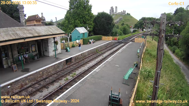 A small train station platform with a stone building on the left, green benches, and a few people sitting or standing. The platform is lined with a yellow fence on the right, beyond which there is a grassy area with trees and a pathway. Overhead, there are electrical lines supported by a wooden pole. In the background, a green hill with a ruined castle on top is visible, and there are railway tracks running through the scene. The timestamp indicates the photo was taken on June 29, 2025, at 1:00 pm.