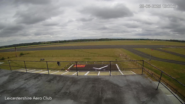 A view from a webcam at Leicestershire Aero Club showing an empty tarmac area with a helicopter landing pad marked with a white 'X' and red and yellow markings. In the background, there is a grassy field, a paved runway, and overcast sky with gray clouds. Two people are visible standing on the grass near the edge of the tarmac. The top right corner displays a timestamp showing "29-06-2025 13:00:31". The bottom left corner shows the label "Leicestershire Aero Club".