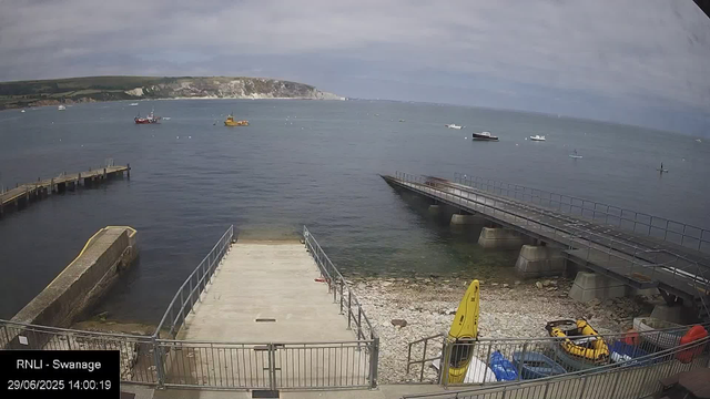 A view of a harbor with calm water, several boats floating in the distance, and a pier extending into the water. In the foreground, there is a small concrete and metal dock area, a yellow caution sign, a yellow inflatable boat, and some equipment and vehicles on a gravel surface. The background shows coastal cliffs and a partly cloudy sky.