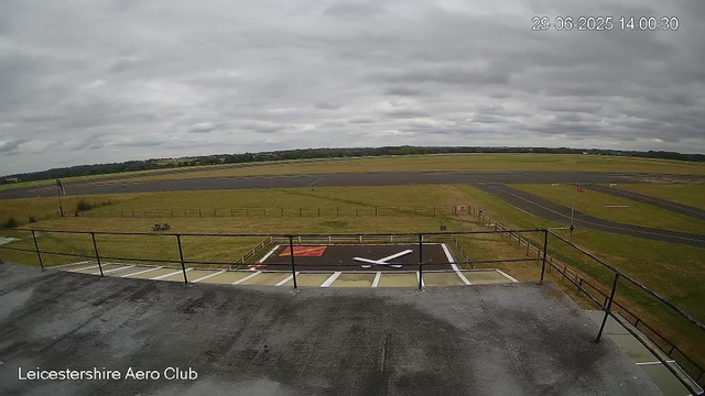An overcast sky covers the runway of Leicester Aero Club. The foreground shows a platform with a railing and a whirlybird mark, while the runway extends into the distance with grassy areas and taxiways on either side. The scene appears to be captured from an elevated structure, possibly a control tower or observation deck. The timestamp in the upper right corner reads "29-06-2025 14:00:30."