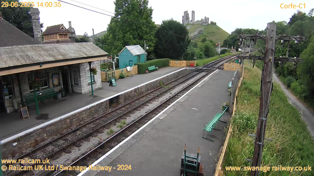 A small railway station platform with a stone building on the left, featuring a shelter, hanging flower baskets, benches, and a signboard. There is a green fence and a small teal-colored shed behind the platform. To the right, there is a wooden pole with electrical wires and a paved path running parallel to the tracks. In the background, a grassy hillside with ruins or castle remains perched at the top is visible, along with lush green trees and a partly cloudy sky. The timestamp indicates it is 14:00 on June 29, 2025.