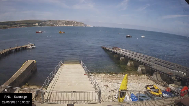 A view of a harbor with several boats floating on calm water, a dock extending into the sea, and a concrete platform with railings in the foreground. There are some yellow safety equipment and small vehicles parked near the platform. In the background, there are green rolling hills and white cliffs under a partly cloudy sky.