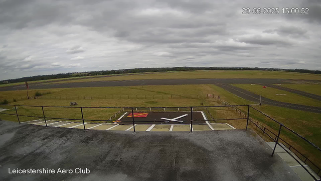 The image shows a view from a webcam at Leicestershire Aero Club on a cloudy day. In the foreground, there is a parking area with a large white X painted on a black surface, possibly a helipad or aircraft landing zone, with a red and yellow marking nearby. A black metal railing surrounds this area, and rain droplets are visible on the camera lens. Beyond, there is a grassy field with a runway in the distance, set under a sky filled with thick, grey clouds. The timestamp indicates the date and time as June 29, 2025, at 15:00:52.