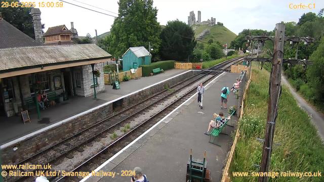 A small train station platform with a stone building on the left side and a row of benches along the right side, where several people are seated. Two people are walking along the platform, one in a gray jacket and the other in a turquoise backpack. A few more people are sitting on benches farther down the platform. Behind the platform, there is a green hut, a park with hedges, and a castle on a hill in the background. A large power pole with wires is on the right side, with a narrow footpath running alongside it. The overall scene appears to be in a rural or semi-rural area with lush green trees and grass around. The sky is mostly cloudy. The time and date are displayed at the top left corner.