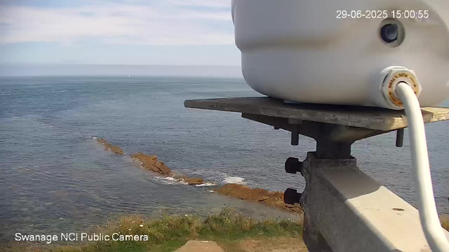 A webcam view of a coastal scene showing the sea with gentle waves and rocky formations near the shore. The sky is partly cloudy with patches of blue. In the foreground, part of a weather instrument or device is mounted on a wooden platform. The image is timestamped "29-06-2025 15:00:55" in the top right corner, and the bottom left corner notes "Swanage NCI Public Camera."