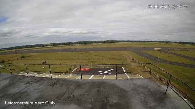 An outdoor view of an airfield shows a cloudy sky with patches of blue. In the foreground, there is a helipad with a white 'H' marking, surrounded by a small fenced area. A few people and a motorcycle are near the helipad. Further back, a small yellow aircraft is parked on a runway. The landscape is flat with grassy areas and a few trees in the distance. The timestamp indicates the date and time as 29th June 2025 at 16:00:04, and the location is Leicesterhire Aero Club.