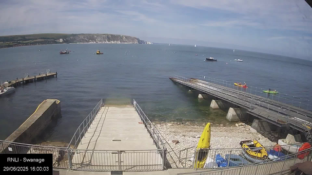A view of a small harbor with a dock extending into calm water, several boats and kayaks anchored, and a rocky beach area with safety equipment including yellow life jackets and a kayak. In the background, the sea stretches out to a distant cliff-lined coast under a partly cloudy sky.