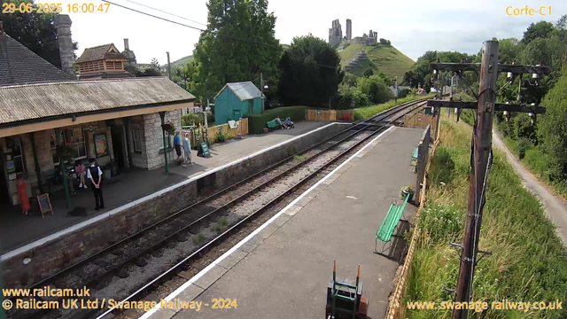 A small train station platform with a stone building on the left, where a few people are waiting and standing near the entrance. There are green benches, a small turquoise shed, and some potted plants along the platform. Railway tracks run parallel to the platform, with overhead electric lines supported by wooden poles. In the background, a green hillside with a ruined castle on top is visible, along with some trees and a cloudy sky. The timestamp shows "29-06-2025 16:00:47," and there are website links and railway credits at the bottom.
