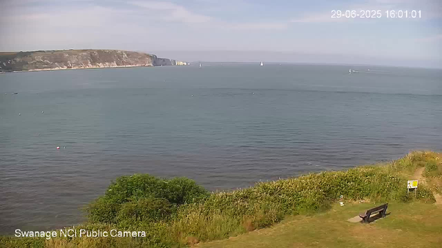A wide view of a calm bay with sailboats on the water and a distant headland with white chalk cliffs under a partly cloudy sky. In the foreground, there is a grassy area with a bench and some bushes. The timestamp in the top right corner shows "29-06-2025 16:01:01".