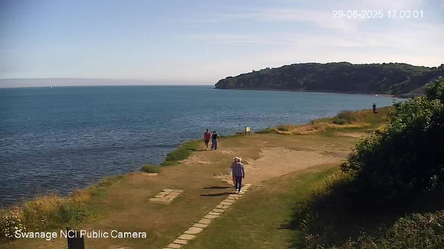 A view of a coastal scene on a sunny day with a person walking along a stone pathway on the grassy shoreline. In the distance, three more people are standing near the water's edge, and another person is further along the shore. The ocean is calm and expansive, with a sailboat visible on the horizon. A large hill covered with greenery rises in the background to the right, and the sky is mostly clear with a few wispy clouds. The timestamp in the top right corner indicates the date and time as June 29, 2025, at 5:00 PM.