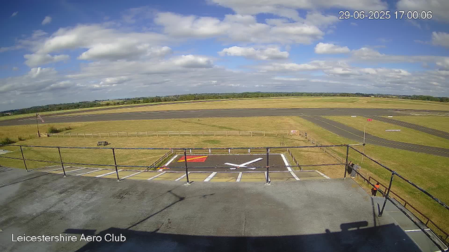 A view from a webcam at Leicesterhire Aero Club shows a runway under a partly cloudy sky. In the foreground, there are marked parking spots for aircraft, including a marked helipad area with a white helicopter symbol. There are a few people near the railing on the right side of the image. The landscape beyond the runway consists of open grassy fields, and the horizon shows a line of trees in the distance. The date and time displayed in the upper right corner are 29-06-2025, 17:00:06.