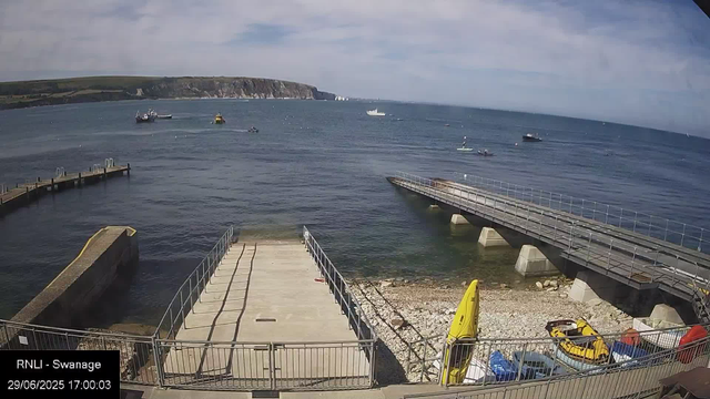 A view of a seaside dock with boats on the water. In the foreground, there is a concrete area with metal railings, a yellow kayak stand, and some parked yellow and red pedal boats. A breakwater extends into the water on the right side, with a small boat nearby. Several boats are scattered in the distance on the calm ocean, with a cliff in the background under a partly cloudy sky.
