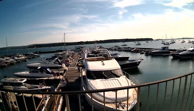 A marina filled with boats and yachts docked along wooden piers. The water is calm, and several sailboats with masts are visible in the distance. The sky is partly cloudy with patches of sunlight. In the foreground, a large white yacht is tied to the dock, and a curved metal railing runs across the bottom of the image. The horizon shows land with some structures, and the overall scene hints at a peaceful, sunny day at the harbor.