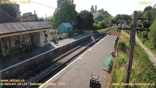 A small train station platform with a stone building on the left, two people sitting on a bench, and a small turquoise shed in the background. The set of railway tracks runs through the middle of the image, curving into the distance. Behind the tracks, there are lush green trees and a hillside with the ruins of an old castle on top. To the right, a tall wooden utility pole with electrical wires and insulators stands beside an overgrown grassy area with a narrow path running parallel. The sky is clear and sunny.