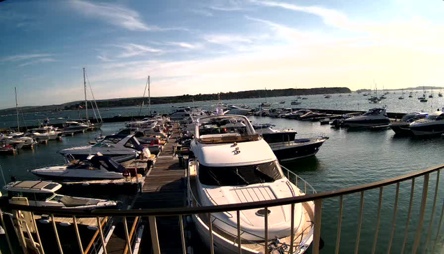 A view of a marina with multiple sailboats and motorboats docked along wooden piers. The water is calm, and the sky shows a mix of clouds and clear areas, with the sun setting in the distance. A curved railing is in the foreground, framing the scene.