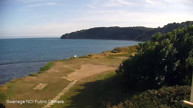 A scenic view of a coastal landscape showing a grassy and sandy shoreline leading to calm blue waters. In the distance, there is a large headland or promontory covered with trees and vegetation. The sky is mostly clear with some wispy clouds, and the image appears to be taken in the late afternoon or early evening. The timestamp in the top right corner reads "29-06-2023 18:09:02."