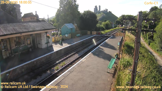 A small railway station platform bathed in sunlight with a building on the left side. The platform has a green bench, a small blue building, and potted plants. Railway tracks run through the scene, with a utility pole and overhead electric lines on the right. In the background, there are trees, some houses, and in the distant background, a castle ruins atop a hill. The date and time displayed in the top left corner is June 29, 2025, at 6:00 PM. The city or station code in the top right corner reads "Corfe-C1."