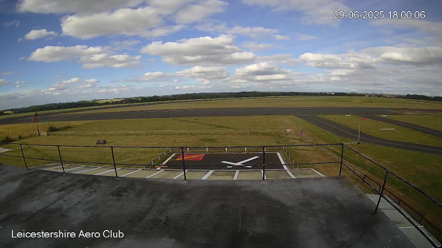 A wide view of an airfield under a partly cloudy sky, with a grassy field, runway, and taxiway extending into the distance. In the foreground, there is a helicopter landing pad marked with a white "H" on a black background, bordered by a railing. The timestamp in the upper right corner reads "29-06-2025 18:00:06," and the label at the bottom left says "Leicestershire Aero Club."