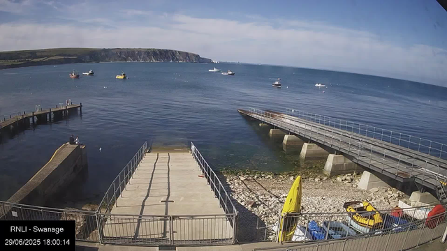 A view of a seaside harbor with boats floating on calm water. There is a concrete pier extending into the water, with a few people sitting on it. A jetty runs parallel to the shoreline, supported by concrete pillars, with a gentle slope leading down from the shore. On the rocky beach area, a yellow caddy and some small vehicles can be seen. In the background, there are cliffs and a partly cloudy sky over the horizon.
