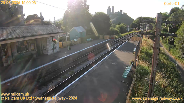 A small railway station platform bathed in sunlight with a building on the left, a small green hut, and benches along the platform. Railway tracks run through the middle, curving off into the distance, with a wooden power pole and overhead electric wires on the right. In the background, there are trees, a hill with ruins or old structures on top, and a clear sky. The image is from a webcam, dated June 29, 2025, at 19:00:05.