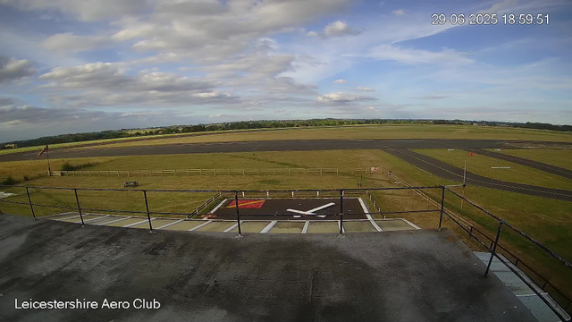 A view of an airfield with runways and open fields under a partly cloudy sky. In the foreground, there is a helicopter landing pad marked with white lines and a red square. The image is captured from a webcam at the Leicesterhire Aero Club on June 29, 2025, at 18:59:51. A fence surrounds the landing area, and there is a small bench near the fence on the grassy area. The horizon shows a distant tree line and open countryside.