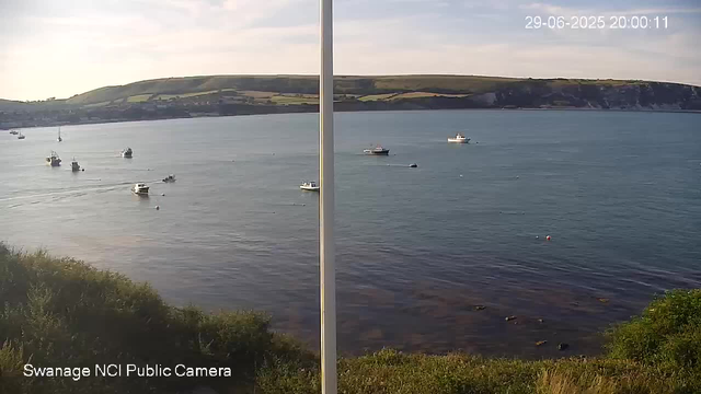 A daytime view of a calm body of water with several small boats scattered across the surface. In the background, there are rolling green hills and a partly cloudy sky. The image is split into two sections by a vertical line in the middle. The bottom left corner has the text "Swanage NCI Public Camera," and the top right corner displays the date and time: 29-06-2025 20:00:11.