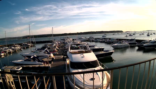 A marina filled with numerous docked boats and yachts extending into calm water, with a bright sky and the sun low on the horizon casting warm light. A curved railing is visible in the foreground.
