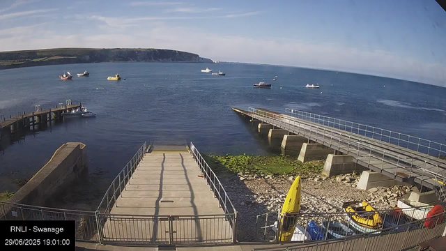 A view of a seaside area with a wooden pier extending into the water, a small boat moored nearby, and several boats scattered across the calm ocean. In the background, there are cliffs and a partly cloudy sky. The foreground shows some parked boats and safety equipment, with a timestamp indicating the date and time as June 29, 2025, at 19:01.