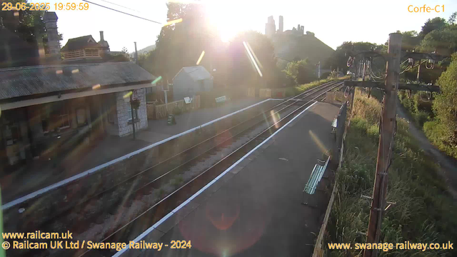 A railway station platform is bathed in bright sunlight with the sun low in the sky, creating sun flares and glare. To the left, there's a small building with a sloped, slightly rusted roof and stone walls. A street lamp is visible near the building. On the right, wooden utility poles with electric wires run parallel to the tracks, and there are bench seats along the platform. In the background, a hillside with a castle or fortress ruins sits atop, surrounded by trees. The atmosphere appears calm and quiet with no trains or people present. The image includes timestamps and website credits at the top and bottom.