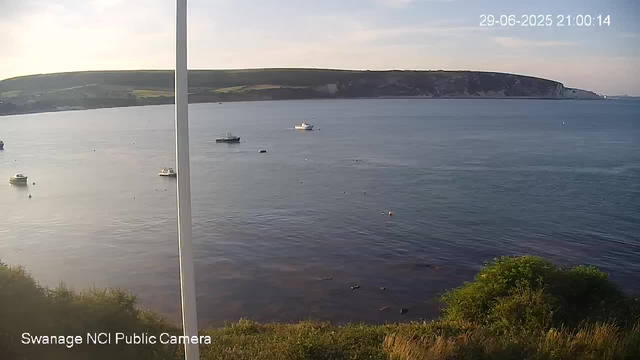 A calm body of water with several small boats floating on the surface. In the background, there is a cliff or shoreline with greenery, and the sky is partly cloudy with soft light indicating late afternoon or early evening. The scene is viewed from a webcam, with date and time displayed in the upper right corner.