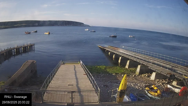 A view of a seaside location with a wooden pier leading into calm water, surrounded by boats anchored in the bay. In the background, there are green cliffs stretching across the horizon under a clear sky. There are some yellow and orange rescue items and safety equipment near the pier. The image is timestamped "29/06/2025 20:00:22" and labeled "RNLI - Swanage."