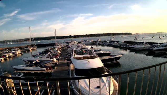 A marina filled with numerous boats and yachts docked along piers, with calm water reflecting the vessels. In the background, there is a clear sky with some clouds, and a distant shoreline with trees. The image appears to be taken during the early evening or late afternoon.