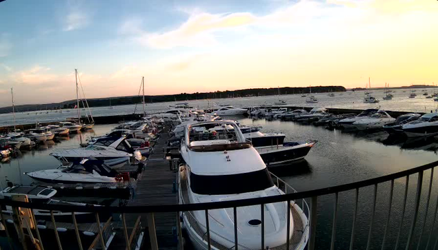 A marina filled with numerous boats and yachts docked along wooden piers, with calm water reflecting the sky. In the background, there are boats sailing on the horizon, and the sky is partly cloudy with a warm, setting sun casting a soft glow on the scene. A black railing curves in the foreground.