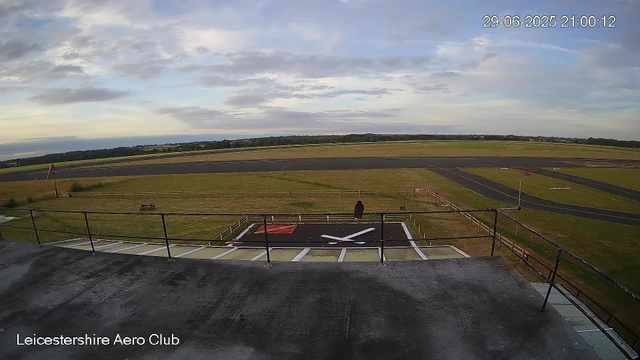 A person stands on a grassy area next to a runway at Leicestershire Aero Club, looking towards the open airfield. The sky is partly cloudy with patches of blue, and the runway extends into the distance. There is a black helicopter landing pad with a white helicopter silhouette painted on it in the foreground. A fence surrounds the area, and there are some signs and markers near the runway. The timestamp on the image shows it was taken on June 29, 2025, at 21:00:12.