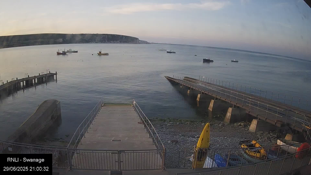 A view of a calm harbor at dusk with several boats anchored in the water. There are two piers extending into the water, one on the right and one on the left. The shoreline is rocky, with some equipment like a yellow kayak and other small boats stored near the water's edge. In the background, there are cliffs along the horizon, and the sky is partly cloudy with soft evening light.