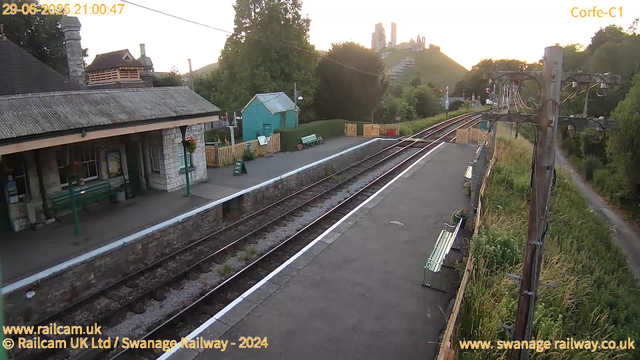 A small train station platform with a stone building on the left, featuring a sloped roof, windows, and flower baskets hanging outside. There are green benches and a sign that says "Way Out." The platform is bordered by a yellow safety line, with a set of train tracks running parallel. On the right, there's a wooden fence with flower pots, tall grass, and trees beyond. In the background, trees, a green shed, some hills, and a distant castle or large building on a hill are visible. The top left corner shows a timestamp and the top right corner displays "Corfe-C1." The sky is mostly clear with the sun setting in the distance.