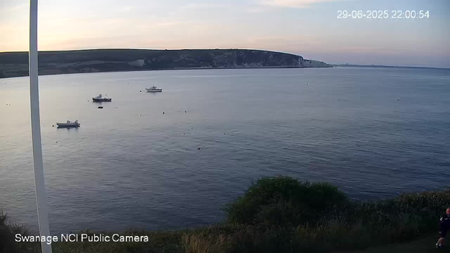 A calm sea with three anchored boats floating near the shoreline. In the background, there are white cliffs stretching across the horizon, and the sky is a soft gradient of light colors, indicating either dawn or dusk. A small section of greenery is visible at the bottom right corner. The timestamp in the top right corner reads "29-06-2025 22:00:54," and the bottom left indicates this is from the "Swanage NCI Public Camera."