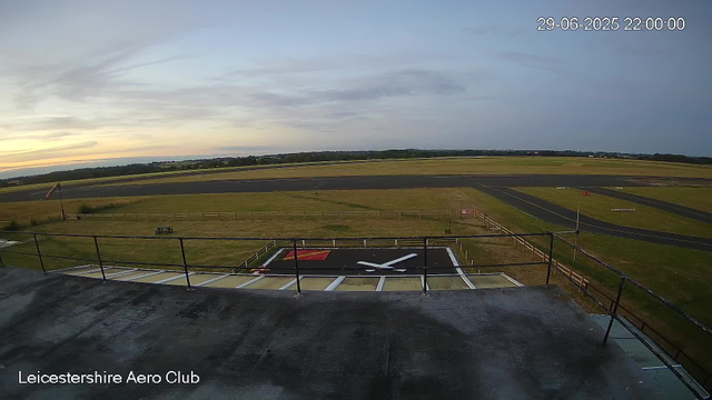 A view from a webcam at LeicesterShire Aero Club showing an empty race track or runway with a large clock painted on the ground in the foreground. The sky is partly cloudy with a hint of sunset colors near the horizon, and the landscape stretches out with fields and distant trees. The timestamp in the upper right corner reads "29-06-2025 22:00:00."