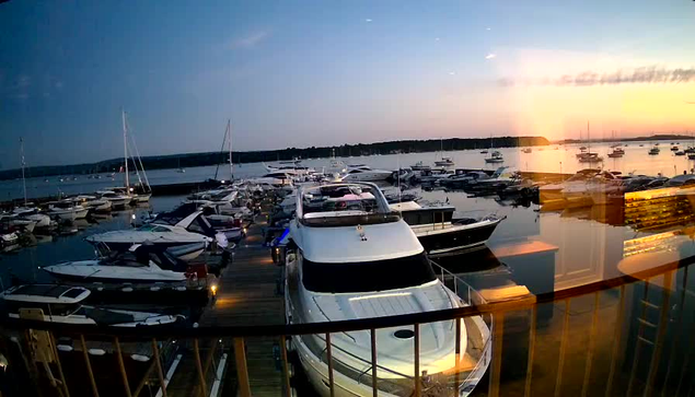 A serene marina at sunset with numerous docked boats and yachts. The sky is a mix of blue and orange hues, reflecting on the calm water. The view is partly obscured by the reflection of indoor lights on a glass window in the foreground.